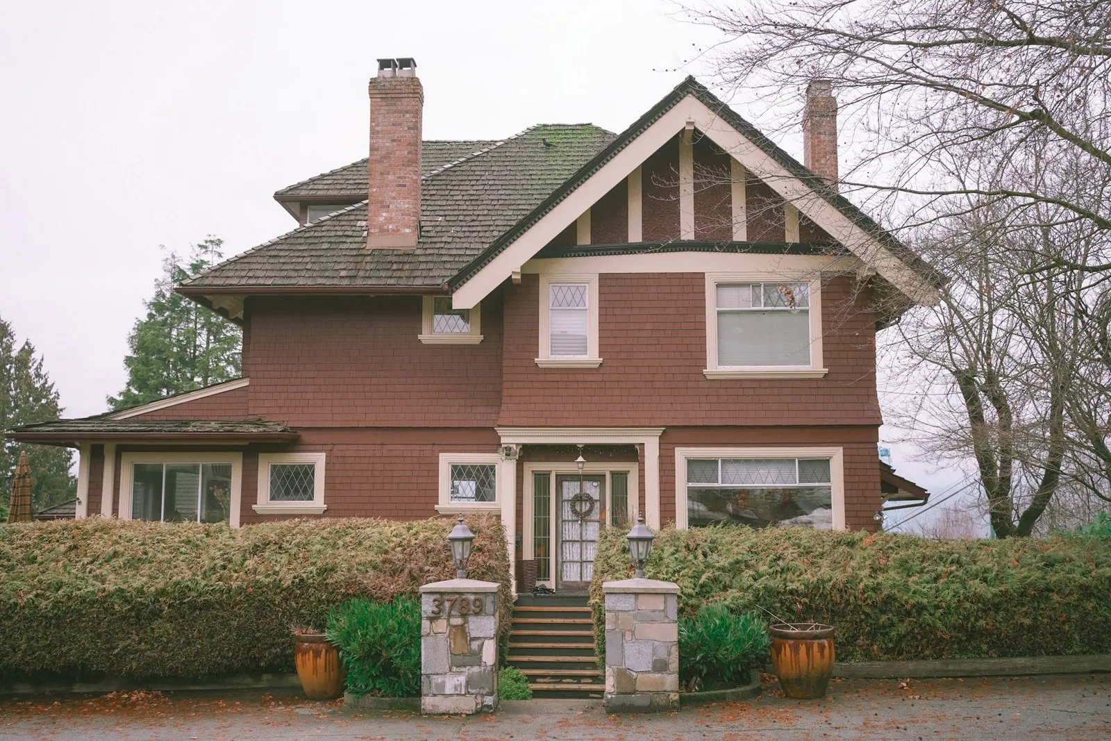 Red-brown Craftsman-era house with brick chimneys and stone gate posts on an overcast day